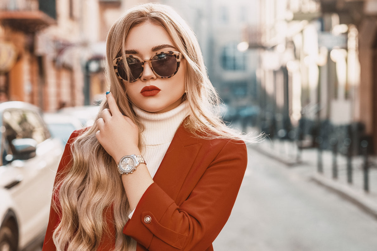A woman in a terracotta blazer and white turtleneck wearing a wrist watch on a city street